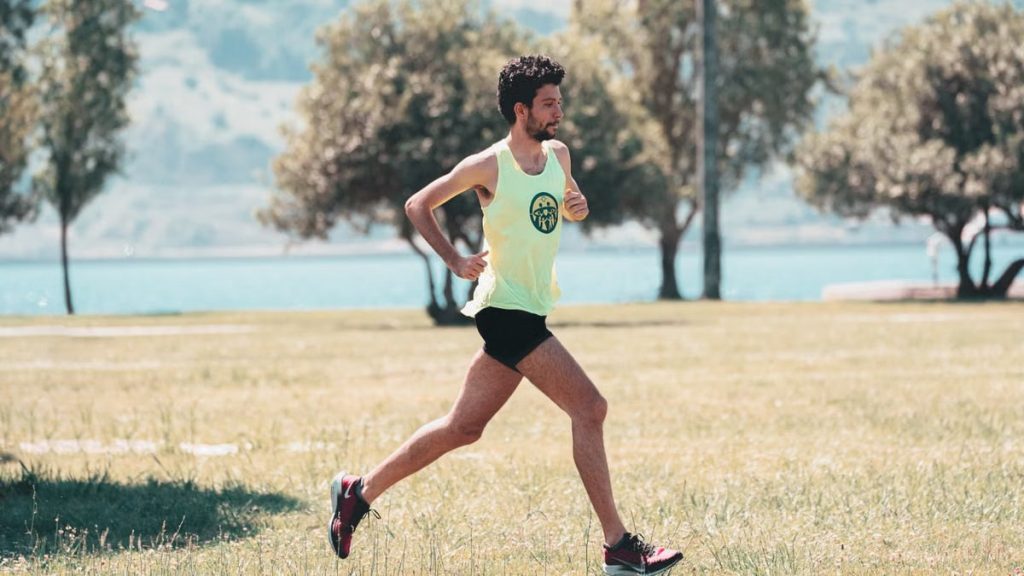 Side view of young determined male athlete in sportswear running on lawn in park near lake in sunny morning