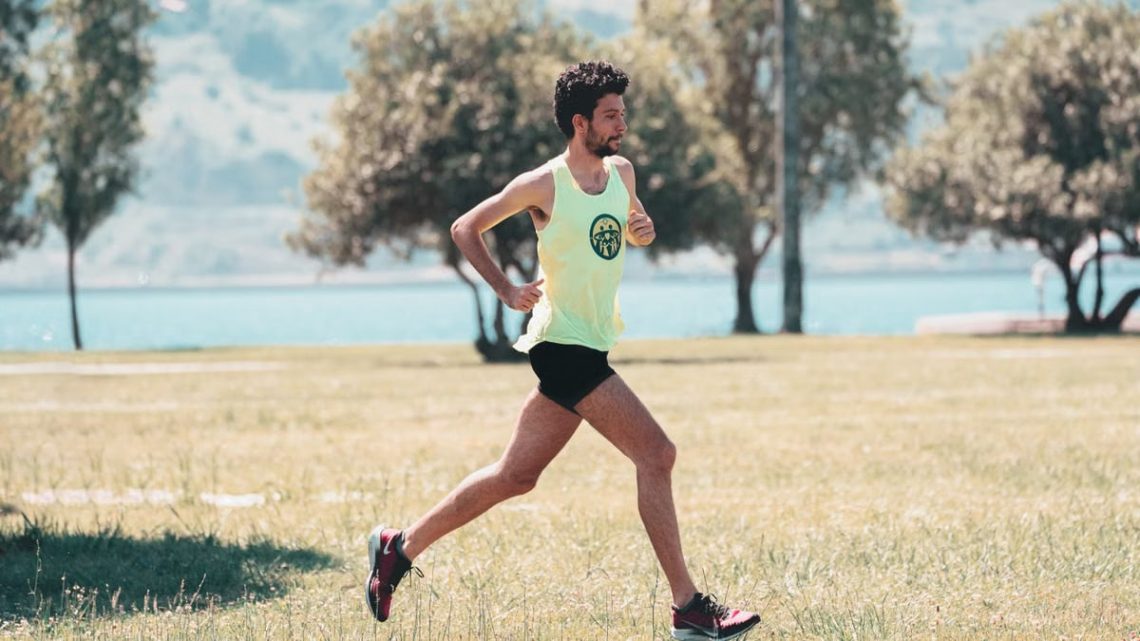 Side view of young determined male athlete in sportswear running on lawn in park near lake in sunny morning